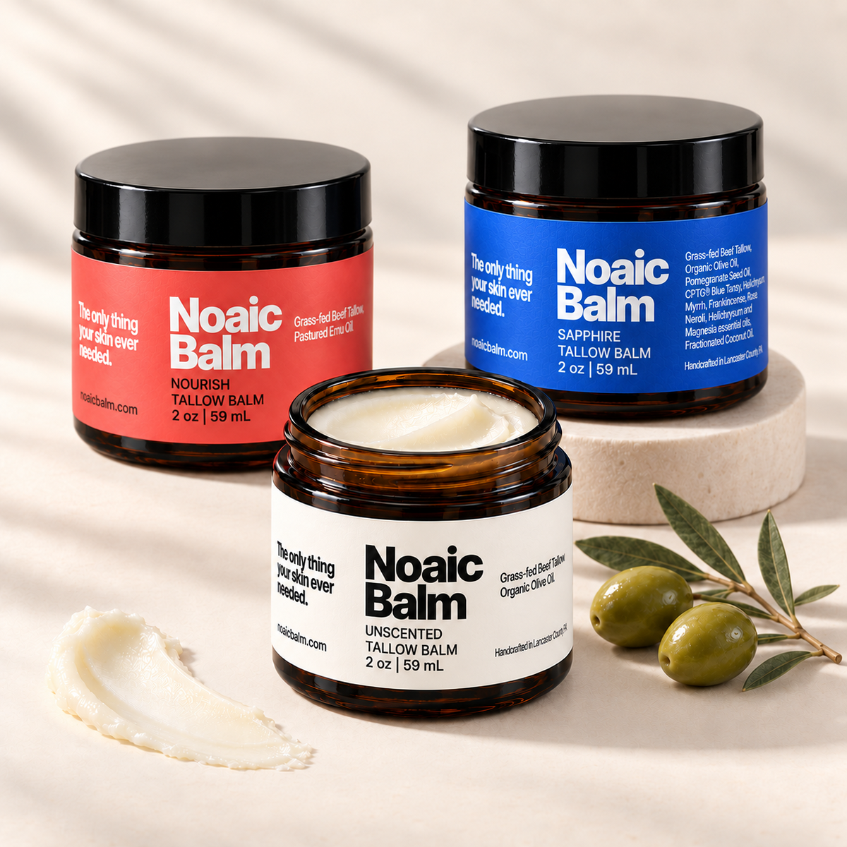 Noaic Balm Nourish, Sapphire, and Unscented tallow balms with olives and a swipe of balm on a cream background.